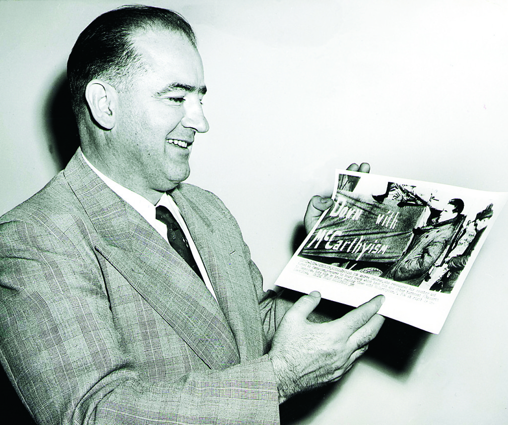 Senator Joseph R McCarthy tittar på ett foto av kommunisten Andrew Condron framför en bil med banderollen"Down with McCarthyism". År 1954. Foto: UPI / TT